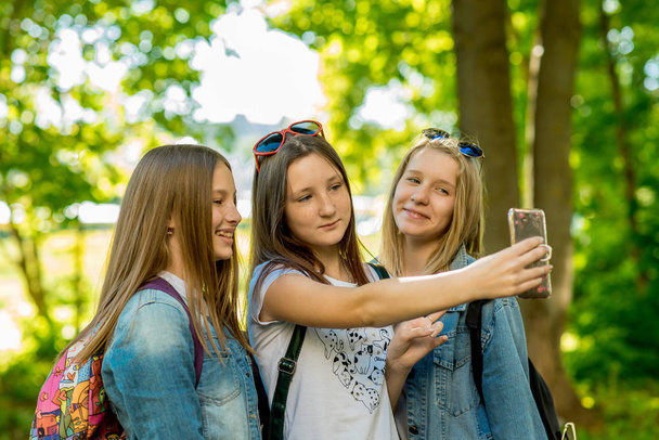 Girls schoolgirls in summer in a park in fresh air. Makes photos pose on the camera. Outdoors in the shade of trees. They are dressed in casual clothes. Pupils are on vacation. - 写真・画像