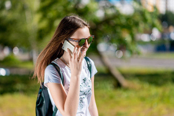 A teenage girl is talking on the phone. In the summer in the park in nature. Brunette in sunglasses. A schoolgirl after school. Emotional happy Smiling in conversation . - Photo, image