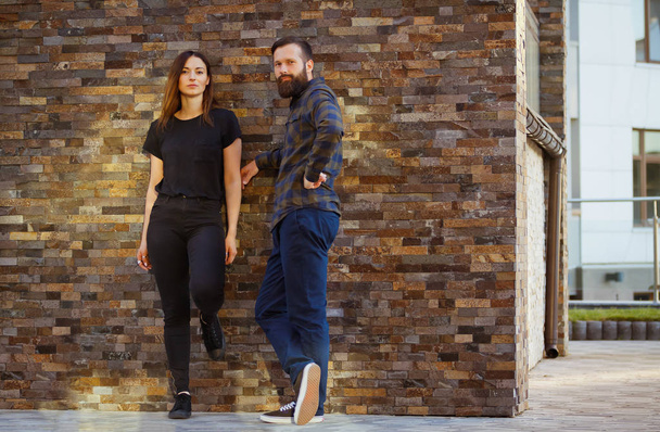 Stylish young couple is spending time together outdoors. Attractive woman and handsome bearded man are posing on brick background. - Foto, Imagen