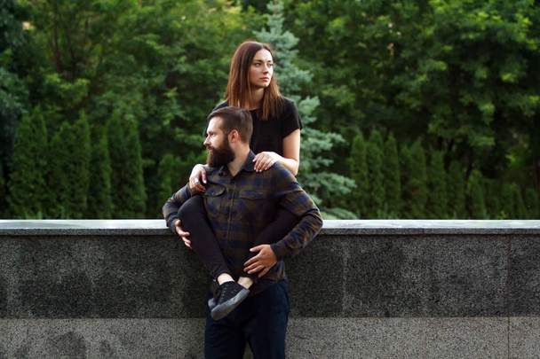 Young couple sitting on parapet in park. Attractive woman and handsome bearded man are standing on street in the city. - Photo, Image