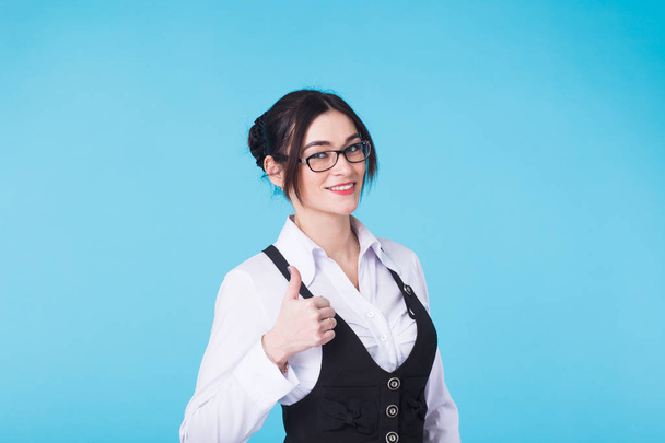 Happy beautiful girl in stylish black glasses showing thumb up symbol on blue background. - 写真・画像