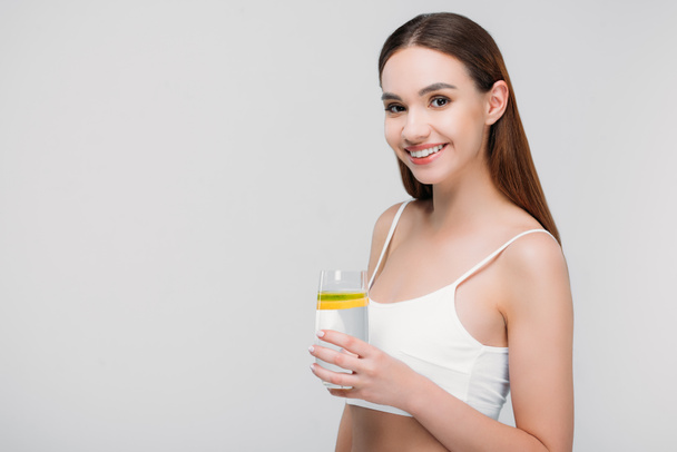 beautiful smiling girl in bra holding lemon water in glass, isolated on grey - Photo, Image