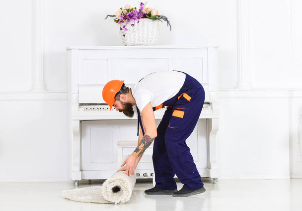 Relocating concept. Courier delivers furniture in case of move out, relocation. Man with beard, worker in overalls and helmet rolling carpet, white background. Loader wrappes carpet into roll - Foto, imagen