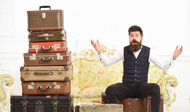 Man, butler with beard and mustache delivers luggage, luxury white interior background. Macho elegant on surprised face sits shocked near pile of vintage suitcase. Luggage and relocation concept - Photo, Image