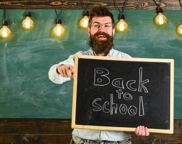 Teacher in eyeglasses holds blackboard with inscription back to school. Back to school concept. Man with beard and mustache on happy face invites students, pointing forward, chalkboard on background - Photo, Image