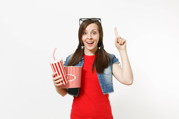 Молодая удивленная женщина в 3D-очках смотрит фильм, держа ведро попкорна, пластиковую чашку соды или колы указывая указательным пальцем вверх на копипространство, изолированное на белом фоне. Эмоции в кино
 - Фото, изображение