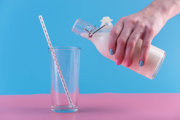 Kobiecej ręki wylewa świeże mleko z butelki szkło do szkła na tle pastel. Kolorowe minimalizm. Pojęcie zdrowe produkty mleczne z wapniem - Zdjęcie, obraz