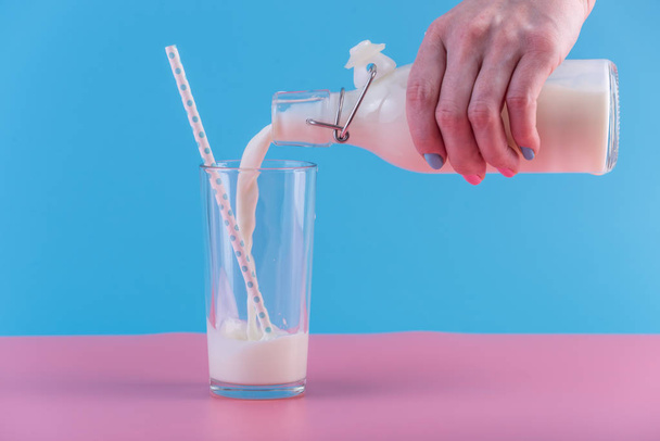 La mano de una mujer vierte leche fresca de una botella de vidrio en un vaso sobre un fondo pastel. Minimalismo colorido. El concepto de productos lácteos saludables con calcio
 - Foto, Imagen