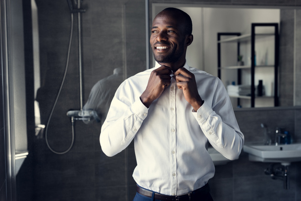 smiling businessman buttoning his white shirt at bathroom and looking away - Photo, Image