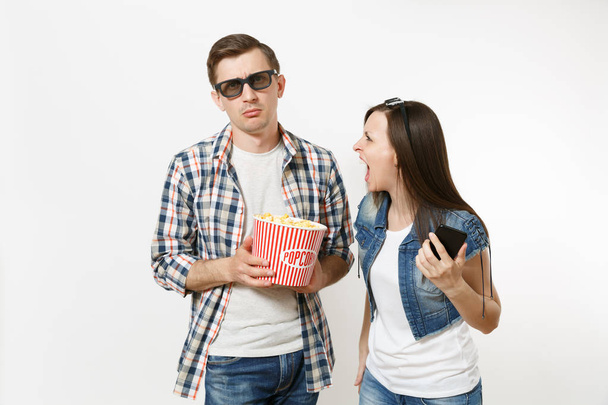 Young irritated couple, woman and man in 3d glasses, casual clothes watching movie film on date holding bucket of popcorn using mobile phone isolated on white background. Emotions in cinema concept - Foto, Bild