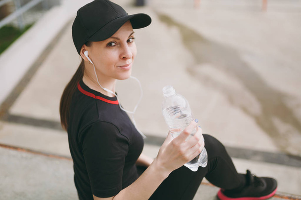 Молодая спортивная улыбающаяся девушка в черной форме, кепка с наушниками, слушая музыку, держа бутылку с водой сидя до или после бега, тренировки в городском парке на открытом воздухе. Фитнес, здоровый образ жизни
 - Фото, изображение