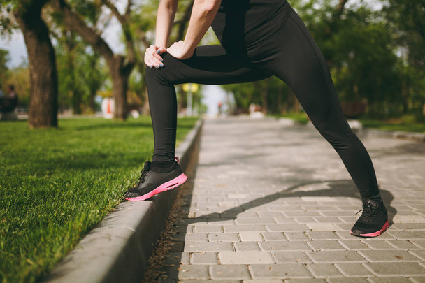 Cropped Young athletic girl in black uniform doing sport stretching exercises, warm-up before running or training, standing in city park outdoors. Fitness, healthy lifestyle concept - Photo, Image