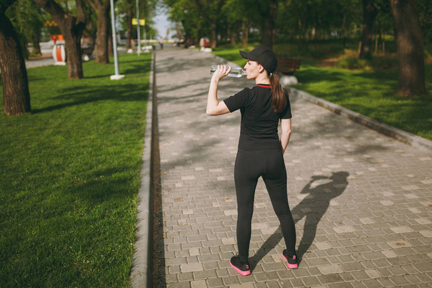 Młoda lekkoatletycznego piękny Dziewczyna Brunetka w czarny mundur i cap gospodarstwa butelka, wody pitnej, podczas treningu przed uruchomieniem stoi w parku na świeżym powietrzu. Fitness, pojęcie zdrowego stylu życia - Zdjęcie, obraz