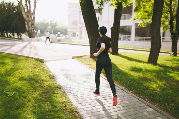 Задний вид Юная спортивная брюнетка в черной форме и кепке тренировки, делать спортивные упражнения и бег, глядя прямо на тропу в городском парке на открытом воздухе. Фитнес, концепция здорового образа жизни
 - Фото, изображение