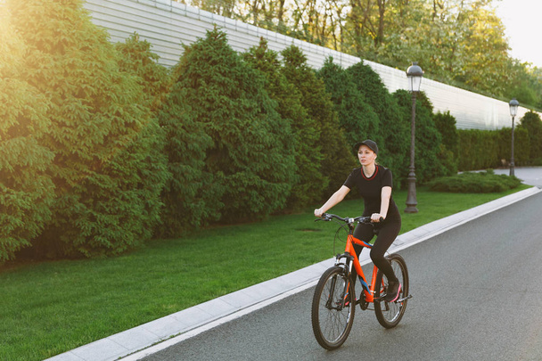 Jeune brune sportive souriante femme forte en uniforme noir, chapeau de route sur vélo noir avec des éléments orange à l'extérieur le printemps ou l'été journée ensoleillée. Fitness, sport, mode de vie sain concept
 - Photo, image