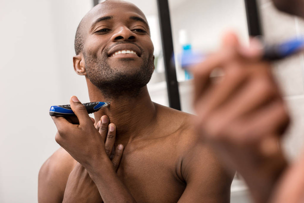 usměvavý mladý muž holení vousů s holicí strojek při pohledu na zrcadlo v koupelně - Fotografie, Obrázek