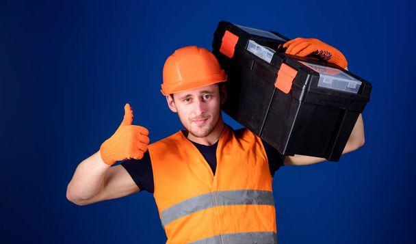Man in helmet, hard hat holds toolbox and shows thumb up gesture, blue background. Worker, repairer, repairman, builder on confident face carries toolbox on shoulder. Repair consultation concept - Foto, imagen