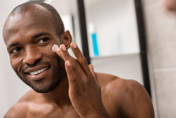 happy young man applying facial cream while looking at mirror in bathroom - Photo, Image
