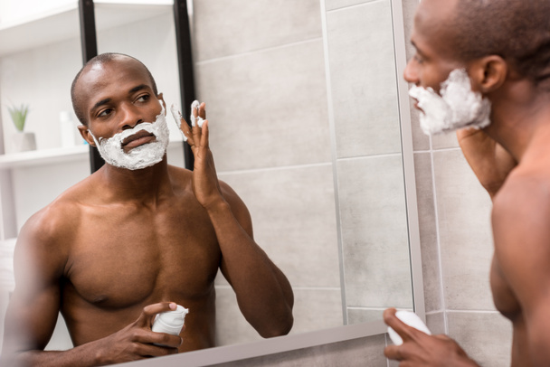 schöner junger Mann trägt Rasiergel auf, während er im Badezimmer in den Spiegel schaut - Foto, Bild