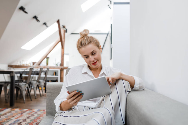 Pretty blonde smiling Caucasian woman sitting on couch at home and typing on tablet. - Foto, Imagem