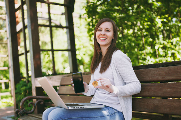 Female showing on camera mobile phone with blank empty screen to copy space. Woman sitting on bench holding bitcoin, modern laptop pc computer outdoors. Mobile Office, online virtual currency concept - Photo, Image