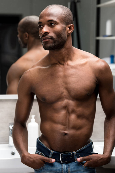 handsome shirtless african american man standing with hands in pockets and looking away in bathroom - Zdjęcie, obraz