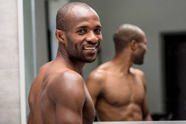 przystojny shirtless african american american człowiek uśmiecha się do kamery, stojąc w łazience - Zdjęcie, obraz
