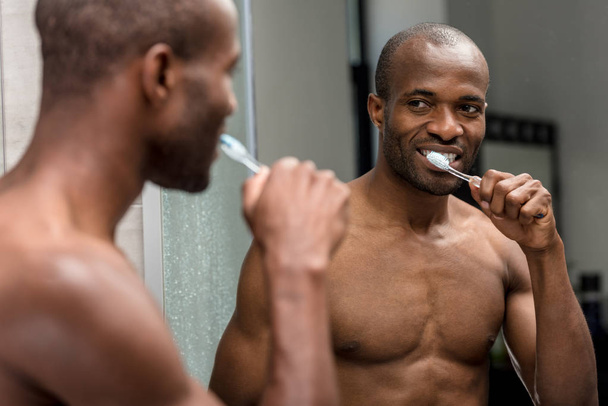 Selektivní fokus usměvavá shirtless afroamerické muže, čistit zuby a při pohledu na zrcadlo  - Fotografie, Obrázek
