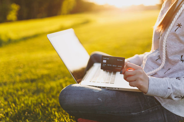 Young happy student female with credit card. Woman sitting on grass ground, working on laptop pc computer in city park on green grass sunshine lawn outdoors. Mobile Office. Freelance business concept - Photo, Image