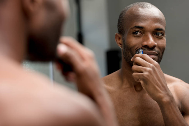selective focus of african american man shaving with electric trimmer in bathroom - Foto, imagen
