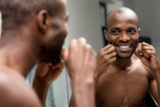若いアフリカ系アメリカ人歯科用フロスを押しながら浴室の鏡を見て笑みを浮かべてください。 - 写真・画像
