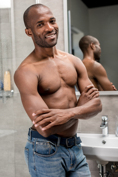 handsome shirtless african american man standing with crossed arms and smiling at camera in bathroom  - Valokuva, kuva