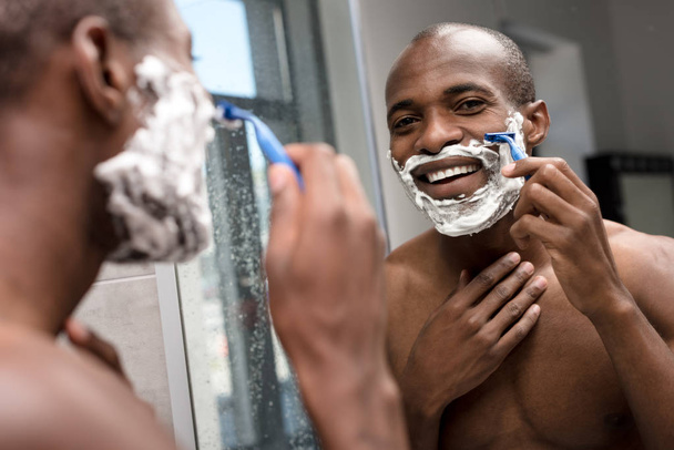 handsome smiling african american man shaving and looking at mirror - Valokuva, kuva
