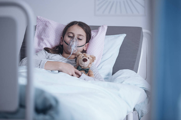 Sick girl with oxygen mask sleeping in a hospital bed with teddy bear - Fotó, kép