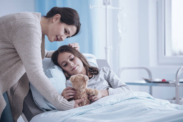 Caring mother giving a plush toy to sick daughter lying in a hospital bed - Photo, image