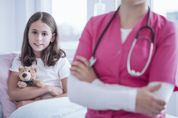 Smiling girl with plush toy during stay in the clinic. Blurred caregiver wearing pink uniform in the foreground - Foto, afbeelding