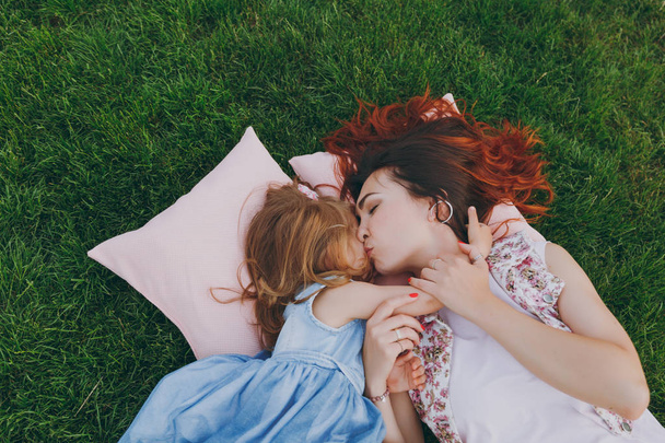 Mujer bonita y niña linda bebé se acuestan en almohadas en la hierba en el parque beso y abrazo, abrazo divertirse. Madre, hija pequeña. Día de la Madre, familia del amor, paternidad, concepto de la infancia
 - Foto, imagen