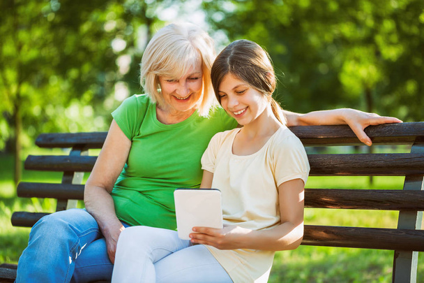 Babcia i wnuczka są siedzi w parku i przy użyciu cyfrowego tabletu.  - Zdjęcie, obraz