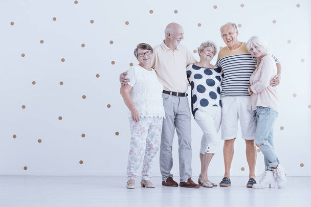 Group of elderly friends hugging each other in white studio with gold dots - Foto, afbeelding