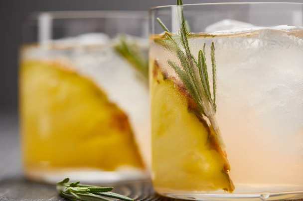 selective focus of two glasses of lemonade with pineapple pieces, ice cubes and rosemary on grey wooden tabletop - Foto, imagen