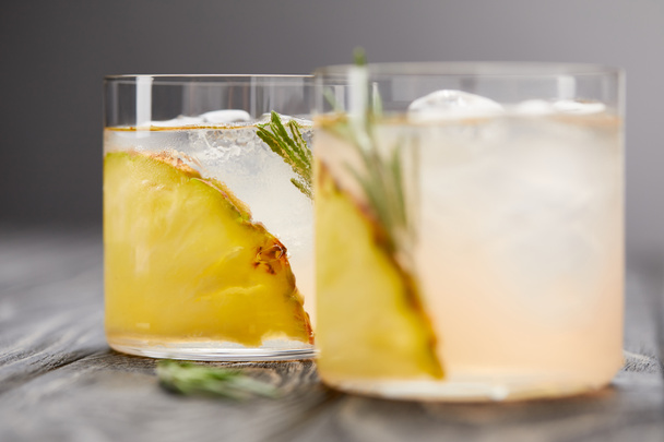 two glasses of lemonade with pineapple pieces, ice cubes and rosemary on grey wooden tabletop - Foto, imagen