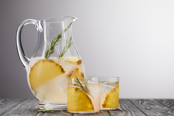 jug and two glasses of lemonade with pineapple pieces, ice cubes and rosemary on grey wooden tabletop - Foto, afbeelding