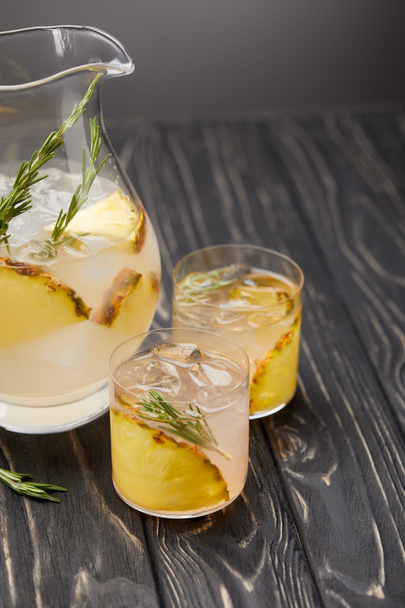 elevated view of jug and two glasses of lemonade with pineapple pieces, ice cubes and rosemary on grey wooden tabletop - Фото, изображение