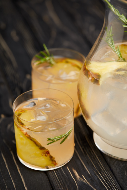 partial view of jug and two glasses of lemonade with pineapple pieces, ice cubes and rosemary on grey wooden tabletop - Foto, afbeelding