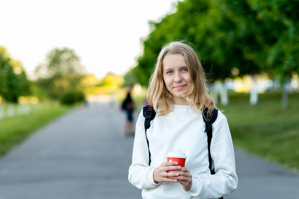 Beautiful girl student blonde. Schoolgirl in summer outdoors. He holds a glass of hot coffee or tea his hand. Smiling happy posing on camera. - Foto, Bild