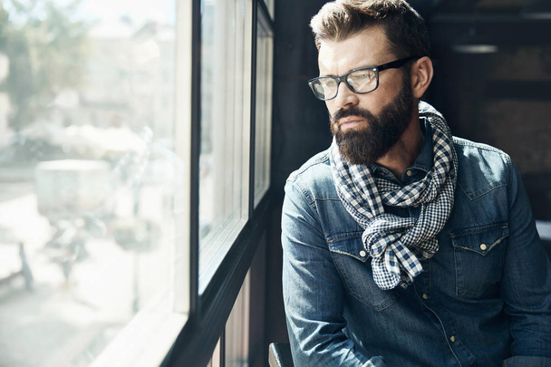 Pensive young man with dark hair and beard, weared in denim jacket, scarf and eyeglasses is sitting near big window  - Zdjęcie, obraz