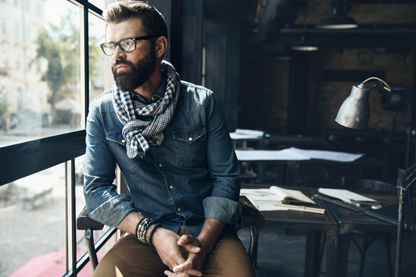 Man architect with dark hair and beard, weared in denim jacket, scarf and glasses is sitting in the modern office near the window with architectural plan on the table behind him - Photo, image