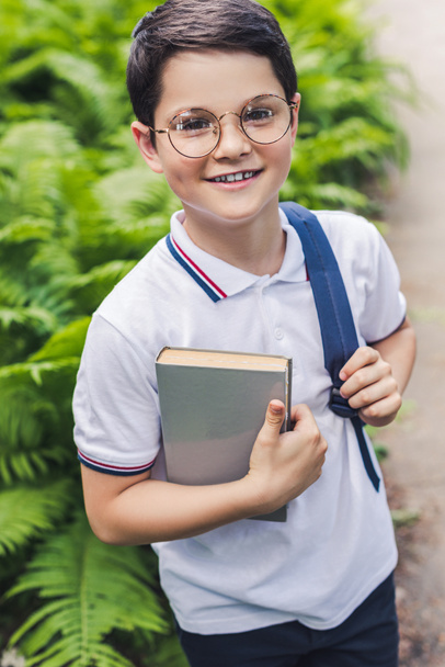 Счастливый школьник с рюкзаком и книгой смотрит на камеру в саду
 - Фото, изображение