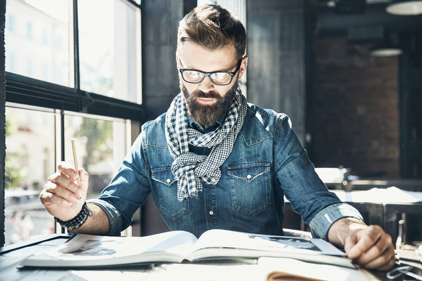 Man architect with dark hair and beard, weared in denim jacket, scarf and eyeglasses is sitting in the modern office near the window and designs a new project - Fotoğraf, Görsel
