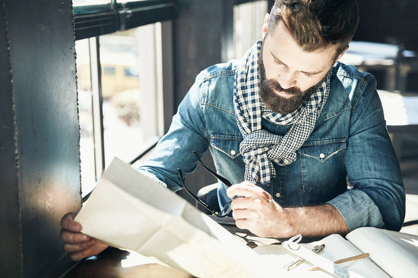 Serious man architect with dark hair and beard, weared in denim jacket, scarf is sitting in the modern office near the window and designs a new project - Photo, image